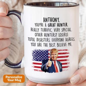 May include: A white ceramic mug with a dark blue handle and interior rim. The mug features a printed American flag background with an image of Donald Trump giving a thumbs up, accompanied by the text: ANTHONY, YOU'RE A GREAT HUNTER. REALLY TERRIFIC, VERY SPECIAL. OTHER HUNTERS? LOSERS! TOTAL DISASTERS. EVERYONE AGREES. YOU ARE THE BEST. BELIEVE ME.
