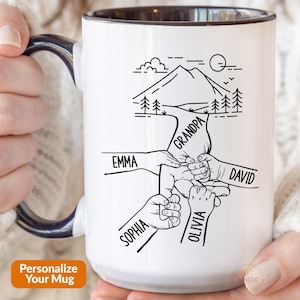 Peut inclure: Un mug en céramique blanche avec une anse et un bord intérieur bleu foncé. L'extérieur présente une illustration au trait noir d'un paysage de montagne au-dessus de plusieurs mains qui se tiennent, avec les noms Emma, Grandpa, David, Sophia et Olivia.
