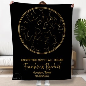 May include: Black blanket featuring a gold constellation design within a circular frame. The text "UNDER THIS SKY IT ALL BEGAN" is above the names "Frankie & Rachel", location, and date. The blanket is held up by a person.