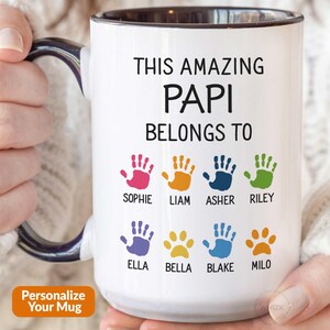 May include: A white ceramic mug with a dark blue interior and handle. The front features the black text THIS AMAZING PAPI BELONGS TO, followed by eight colorful handprints and paw prints, each labeled with names: Sophie, Liam, Asher, Riley, Ella, Bella, Blake, Milo.