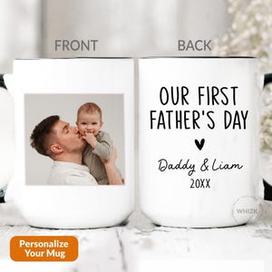 May include: A white ceramic mug with a black handle and rim. The front features a photograph of a person kissing a baby. The back displays the black text "OUR FIRST FATHER'S DAY" above a heart icon, the names "Daddy & Liam", and the year "20XX".