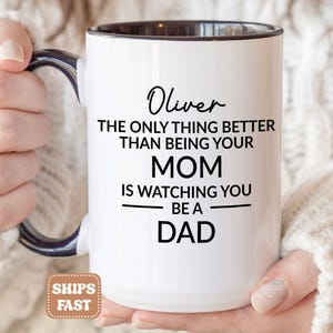 May include: A white ceramic mug with a dark blue interior and handle. The mug features black printed text that reads: Oliver THE ONLY THING BETTER THAN BEING YOUR MOM IS WATCHING YOU BE A DAD. The design is centered on the front of the mug.