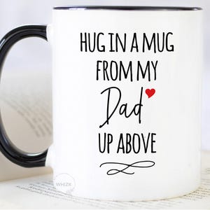 May include: White ceramic mug with a black handle and rim. The mug features the text "HUG IN A MUG FROM MY Dad UP ABOVE" in black script, with a small red heart. A decorative swirl is below the text.