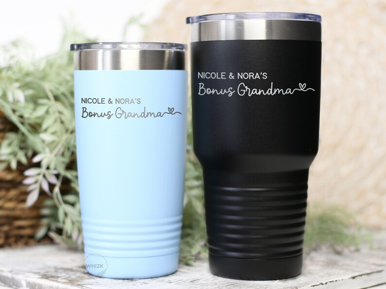 May include: Two stainless steel tumblers in black and light blue. Both tumblers are engraved with the text "Nicole & Nora's Bonus Grandma" and a heart symbol.