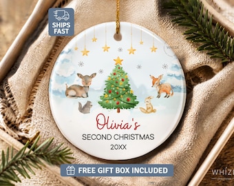 May include: A round white ceramic Christmas ornament featuring a central green Christmas tree surrounded by a deer, a fox, a rabbit, and a squirrel. The text reads Olivia's SECOND CHRISTMAS 20XX. Gold stars hang from the top edge on thin strings.