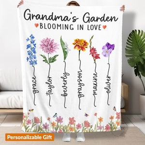 May include: White fleece blanket with a personalized floral design. The text reads "Grandma's Garden Blooming in Love" with a heart symbol on each side of the text.  The blanket features five different flowers with names written underneath each flower: Grace, Taylor, Beverly, Grayson, Maxine, and Oliver.