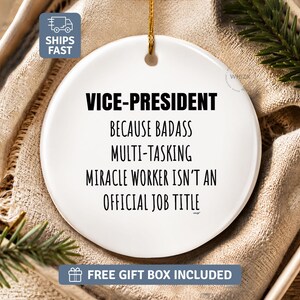 May include: A round white ceramic ornament featuring black text that reads: VICE-PRESIDENT Because Badass Multi-Tasking Miracle Worker Isn't An Official Job Title. The ornament includes a gold hanging string and is presented in a neutral-colored box.