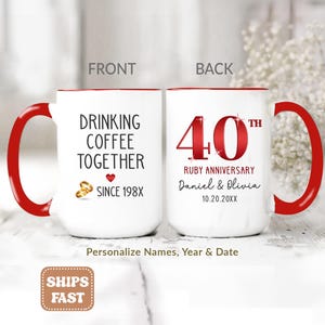 May include: Two white ceramic mugs with red handles and interiors. One mug features the text DRINKING COFFEE TOGETHER SINCE 198X with two gold rings and a red heart. The other mug displays 40TH RUBY ANNIVERSARY with the names Daniel & Olivia.