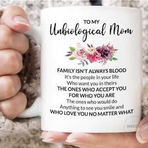 May include: White ceramic mug with the text "TO MY Unbiological Mom" and a floral design. The mug also features a quote about family. The mug is held by a person.