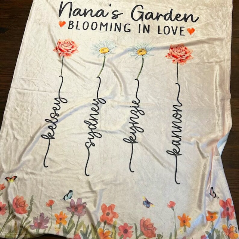May include: A white fleece blanket with the text "Nana's Garden Blooming in Love" and four flowers with names written underneath: Kelsey, Sydney, Kyngzie, and Kaimnon.