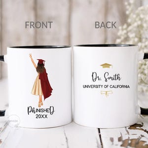 May include: Two white ceramic mugs with black handles and rims. The left mug features a graduation illustration of a person in a maroon gown and cap with the text PHINISHED 20XX. The right mug displays the text Dr. Smith, UNIVERSITY OF CALIFORNIA.