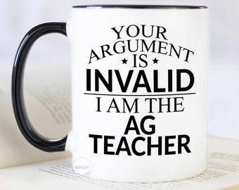 May include: A white ceramic mug with a black handle and rim, featuring black text that reads: YOUR ARGUMENT IS INVALID I AM THE AG TEACHER. The mug has a cylindrical shape and a smooth finish, with the text centered on the front surface in a bold font.
