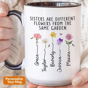 May include: A white ceramic mug with a dark blue handle and interior rim. The mug features the text "SISTERS ARE DIFFERENT FLOWERS FROM THE SAME GARDEN" above five floral illustrations, each with a name written vertically: Grace, Taylor, Beverly, Jessica, and Maxine.