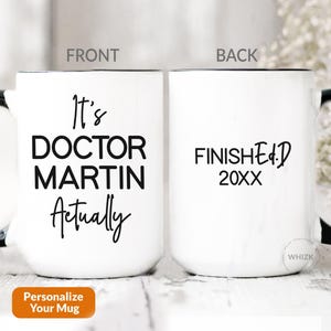 May include: A white ceramic mug with a black handle and interior. The front features the black text It's DOCTOR MARTIN Actually. The back displays the black text FINISHED 20XX. The mug is shown from two angles labeled FRONT and BACK respectively.