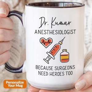 May include: White ceramic coffee mug with a black handle and black rim. The mug has a design that says "Dr. Kumar Anesthesiologist" with a red heart and a syringe. The text below the design says "Because Surgeons Need Heroes Too".