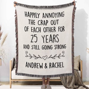 May include: A cream-colored woven throw blanket with black fringe edges, featuring the text "HAPPILY ANNOYING THE CRAP OUT OF EACH OTHER FOR 25 YEARS AND STILL GOING STRONG" above a decorative vine and heart graphic, with the names "ANDREW & RACHEL" below.