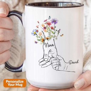 May include: White ceramic mug with a black handle. The mug features a black line drawing of two hands holding each other, with a bouquet of colorful flowers growing from the top of the hands. The text "Nina" and "David" are written on the hands.