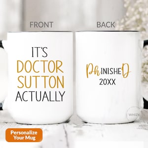 Peut inclure: Un mug en céramique blanche avec une anse et un bord noirs. L'avant affiche le texte IT'S DOCTOR SUTTON ACTUALLY en noir et doré. L'arrière présente le texte PhINISHED 20XX en doré et noir. Le mug est présenté sous deux angles différents.