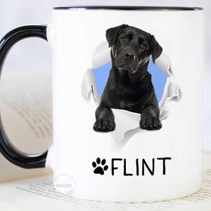 May include: A white ceramic mug with a black handle and rim, featuring a graphic of a black Labrador retriever peeking through a torn paper effect. Below the dog, the name FLINT is printed in black capital letters next to a black paw print icon.