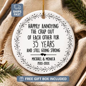 May include: A round white ceramic ornament featuring a black leaf wreath border and the text: Happily annoying the crap out of each other for 35 years and still going strong, Michael & Monica, 19XX-20XX, with two small black hearts.