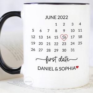 May include: A white ceramic mug with a black handle and rim, featuring a printed June 2022 calendar. The number 16 is circled with a red heart. Below the calendar, the text reads first date followed by the names DANIEL & SOPHIA with a red heart.
