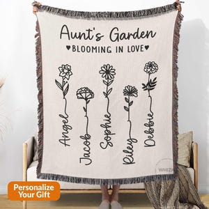 May include: A cream-colored woven throw blanket featuring black text that reads Aunt's Garden and BLOOMING IN LOVE. The design includes five black line-art flower illustrations, each paired with the names Angel, Jacob, Sophie, Riley, and Debbie.