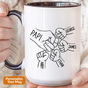 May include: A white ceramic mug with a dark blue handle and interior. The exterior features a black line art graphic of five fists in a group fist bump. The names Papi, George, James, Ethan, and Abby are printed next to each corresponding fist.