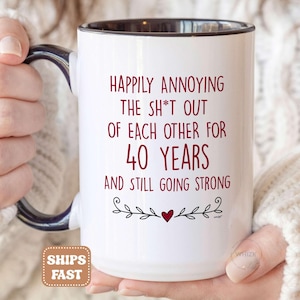 May include: A white ceramic mug with a dark interior and handle, featuring red text that reads: Happily annoying the sh*t out of each other for 40 years and still going strong, finished with a small red heart and black decorative flourishes.