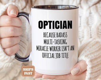 May include: A white ceramic mug with a dark blue interior and handle. The front features black text that reads: OPTICIAN BECAUSE BADASS MULTI-TASKING MIRACLE WORKER ISN'T AN OFFICIAL JOB TITLE. The mug has a cylindrical shape and a smooth finish.