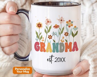 May include: A white ceramic mug with a dark interior and handle, featuring a colorful floral illustration above the word GRANDMA in large, rounded, multi-colored letters, with the text est. 20XX printed in black below the floral design.