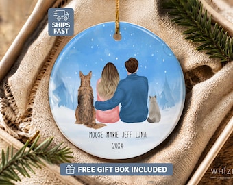 May include: A round ceramic ornament featuring a winter scene with a brown dog, a person with long hair in a pink sweater, a person in a blue sweater, and a grey cat sitting together. The text reads MOOSE MARIE JEFF LUNA and 20XX below them.