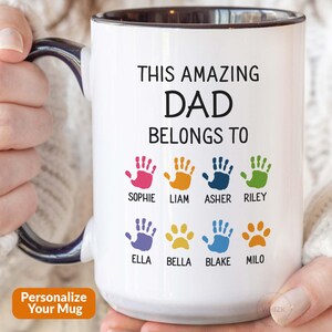 May include: A white ceramic mug with a dark blue handle and interior. The front features the black text THIS AMAZING DAD BELONGS TO above eight colorful handprints and paw prints, each labeled with names including Sophie, Liam, Asher, and Riley.