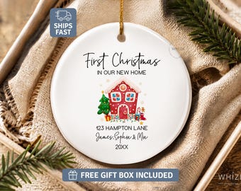 May include: A round white ceramic ornament featuring a red gingerbread house illustration, a green Christmas tree, and text that reads First Christmas IN OUR NEW HOME, 123 HAMPTON LANE, James, Sophia & Mia, and 20XX, suspended by a gold string.