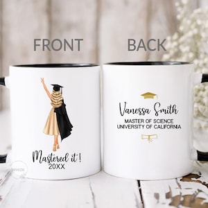 May include: White ceramic coffee mug with a black rim and handle. The front of the mug features a cartoon illustration of a woman in a graduation cap and gown. The text "Mastered it! 20XX" is printed below the illustration. The back of the mug has the text "Vanessa Smith Master of Science University of California" printed in black with a gold graduation cap and diploma illustration.