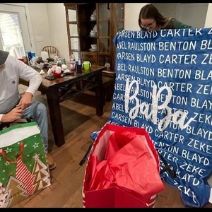 May include: A blue blanket with white text that reads "CARSEN BLAYD CARTEK ZEKE AFEL RAULSTON BENTON BLAYD CARSEN BLAYD CARTER ZEKE BEL RAULSTON BENTON BLAYD CARSEN BLAYD CARTER ZEKE ABEL ALLSTON BENTON BLAYD CARCA TER ZEKE ABELKISTONBY TUN BL CARSEN BLAYD CARTER ZEKE BLAYD ADC ENTON BLAYD RTER ZEKE KAYD CKE". The blanket is folded in half and is being held by a person. There are two gift bags in the foreground, one green and one red.