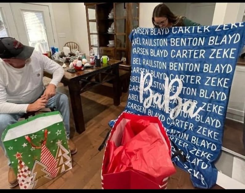 May include: A blue blanket with white text that reads "CARSEN BLAYD CARTEK ZEKE AFEL RAULSTON BENTON BLAYD CARSEN BLAYD CARTER ZEKE BEL RAULSTON BENTON BLAYD CARSEN BLAYD CARTER ZEKE ABEL ALLSTON BENTON BLAYD CARCA TER ZEKE ABELKISTONBY TUN BL CARSEN BLAYD CARTER ZEKE BLAYD ADC ENTON BLAYD RTER ZEKE KAYD CKE". The blanket is folded in half and is being held by a person. There are two gift bags in the foreground, one green and one red.