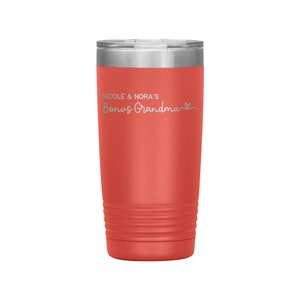 May include: Coral red stainless steel tumbler with a silver lid. The tumbler has a white inscription that reads "Nicole & Nora's Bonus Grandma" with a small heart symbol.