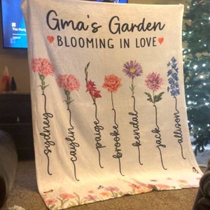 May include: A white fleece blanket with the text "Gma's Garden Blooming in Love" and a floral design with seven flowers and names written underneath each flower. The names are Sydney, Caylin, Paige, Brooke, Kendal, Jack, and Allison.