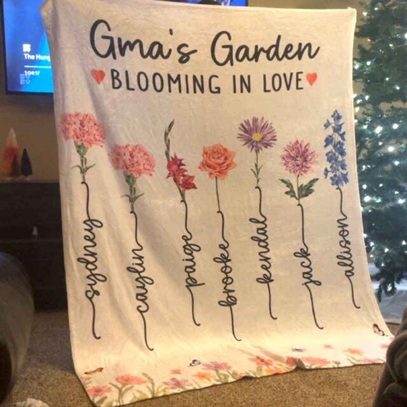 May include: A white fleece blanket with the text "Gma's Garden Blooming in Love" and a floral design with seven flowers and names written underneath each flower. The names are Sydney, Caylin, Paige, Brooke, Kendal, Jack, and Allison.