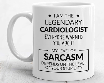 May include: A white ceramic mug with a handle on the left side, featuring black printed text that reads: I AM THE LEGENDARY CARDIOLOGIST EVERYONE WARNED YOU ABOUT. MY LEVEL OF SARCASM DEPENDS ON THE LEVEL OF YOUR STUPIDITY, framed by two black stars.