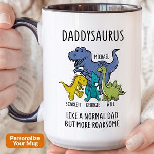 May include: A white ceramic mug with a dark blue handle and interior rim. The mug features a graphic of four dinosaurs with the text DADDYSAURUS, the names Michael, Scarlett, Georgie, and Will, and the text LIKE A NORMAL DAD BUT MORE ROARSOME.