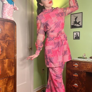 May include: A woman wearing a pink and purple patterned pantsuit with a matching belt. She is standing in a room with a wooden dresser and a rug on the floor.