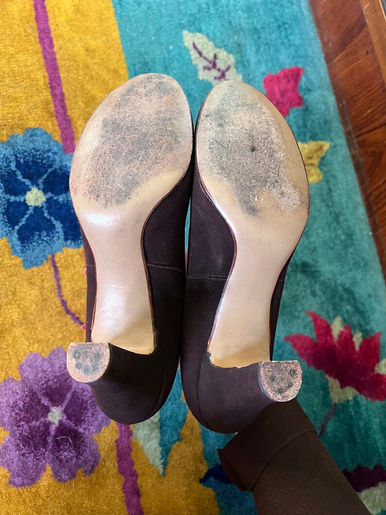 May include: A pair of brown suede heeled shoes with a light brown sole. The shoes are on a patterned rug.