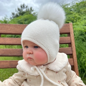 May include: A white knit baby hat with a large, fluffy pom-pom on top. The hat has ear flaps and a chin tie. The baby is wearing a cream-colored outfit with a floral pattern and a ruffled collar. The background is blurred with green foliage.