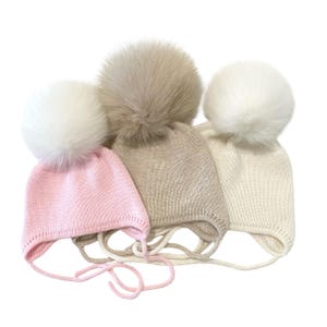 May include: Three knit baby hats with large faux fur pom-poms. The hats are in shades of pink, beige, and cream. Each hat has a chin strap with ties. The hats are designed for infants and toddlers.