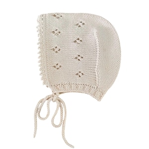 Puede incluir: Un gorro de punto color crema con un delicado patrón de calados en forma de diamante en la parte superior. La prenda incluye un borde acanalado en la parte frontal, un detalle festoneado y dos lazos trenzados largos para atar bajo la barbilla.