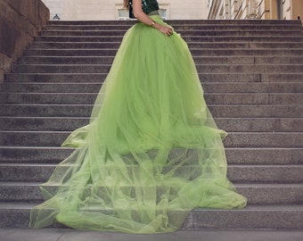 May include: A woman in a green tulle skirt and a sequined top poses on a grand staircase. The skirt is a vibrant, flowing green, contrasting with the darker green top. The background features a large building with multiple windows.