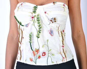 May include: White strapless top with floral embroidery. The bodice features a sweetheart neckline and is adorned with colorful embroidered flowers, green leaves, and small beaded accents. The top is paired with a black skirt.