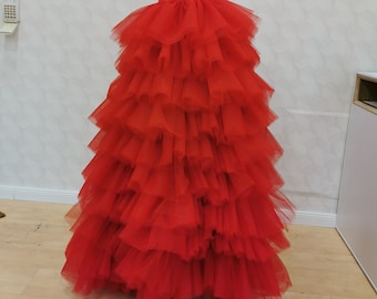 May include: A full-length, tiered red tulle skirt displayed on a black mannequin. The skirt features multiple layers of ruffled fabric, creating a voluminous and textured appearance. The mannequin has a black turtleneck top.