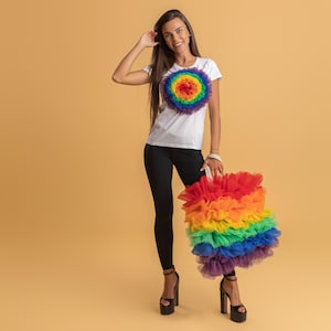 Op de afbeelding: Een wit t-shirt met een regenboogontwerp en een bijpassende regenboog tutu. De persoon draagt ​​een zwarte legging en plateau hakken. De achtergrond is effen beige.